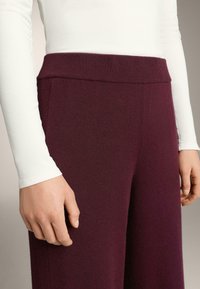 Pantaloni tricotati burgundy cu talie lată; textură moale și croială lejeră. Cămașă albă cu mâneci lungi parțial vizibilă în partea de sus.