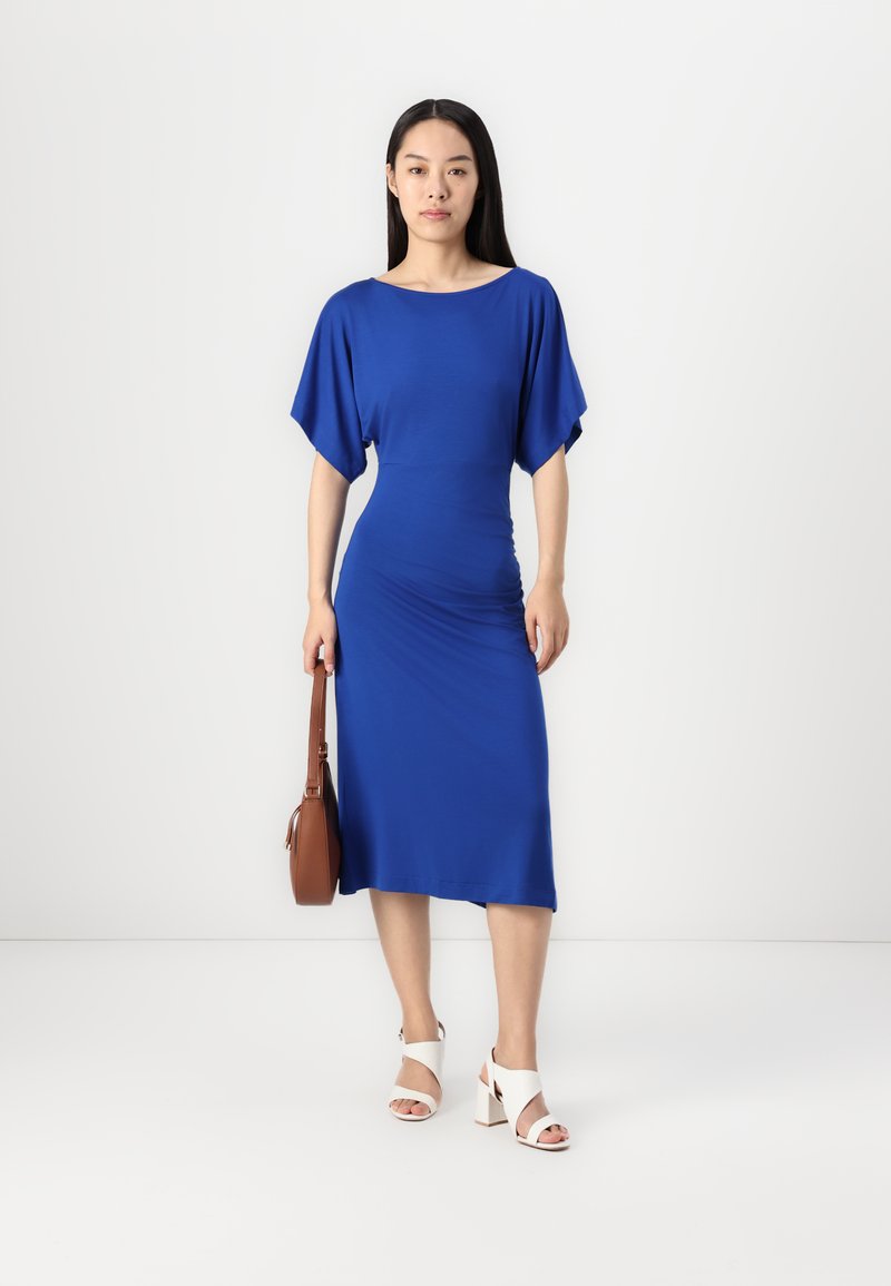 Blaues Midikleid mit Batwing-Ärmeln aus glattem Stoff. Kombiniert mit weißen Absatzsandalen und einer braunen Schultertasche.