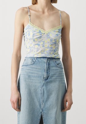 Kobieta ubrana w dopasowany niebiesko-żółty kwiecisty top na ramiączkach z koronkowym wykończeniem, zestawiony z jasnoniebieską, wysokowasową spódnicą jeansową z rozcięciem z przodu.