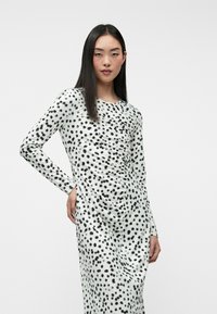 Robe à manches longues en tissu blanc à pois noirs, présentant une silhouette ajustée et des détails froncés à la taille. Texture lisse.