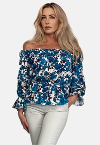 Niebieska bluzka bez ramiączek w kwiaty z marszczonym korpusem, falbanastymi rękawami i białym tłem. W zestawie z jasnymi spodniami.