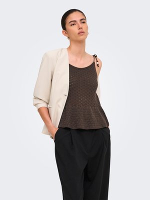 Jeune femme portant un blazer beige sur un top sans manches en maille marron foncé et un pantalon noir, debout les mains dans les poches sur un fond uni.
