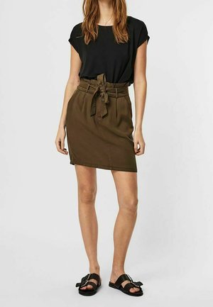 Femme portant un haut noir à manches courtes, une jupe olive taille haute avec une ceinture nouée, et des sandales noires à boucle sur un fond clair et uni.