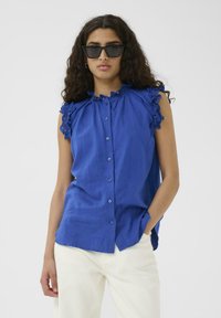 Kék, ujjatlan, gombos ing fodros csipkebetéttel, puha textúrával és laza szabással, fehér nadrággal párosítva.