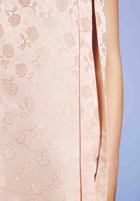 Bešvitrė šviesiai rožinė suknelė su subtiliu gėlių raštu, rodanti šoną plyšį ir nuogą ranką prieš levandų fono.