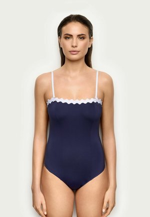 Femme portant un maillot de bain une pièce bleu marine avec un bord festonné blanc et des fines bretelles blanches, debout devant un fond clair uni.