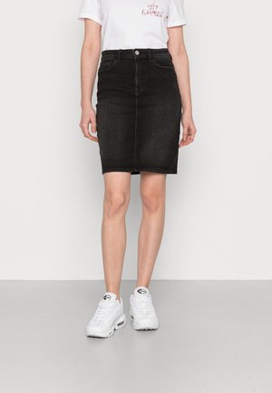 Person iført en sort denimnederdel til knæet, hvid T-shirt og hvide sportssko, stående mod en ensfarvet lys baggrund.