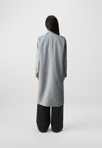 Lys grå lang frakk laget av stoff, med rett snitt, nedbrettbar krage og knappede mansjetter, kombinert med vide svarte bukser.