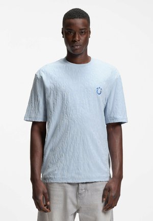 Hombre con camiseta azul claro texturizada con un pequeño logo azul "H2O" en el pecho, vistiendo pantalones gris claro, de pie contra un fondo blanco.