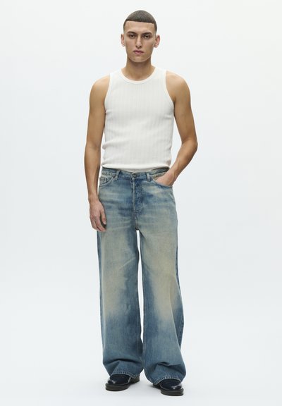 Vide ben, falmet blå denimjeans med lys vask, fem lomme-design og lett slitasje, kombinert med en hvit ribbestrikket singlet.