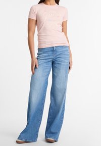 Růžové priléhavé tričko s logem "Guess California", spárované se světle modrými širokými džíny. Tričko je měkké, džíny mají vybledlou texturu.