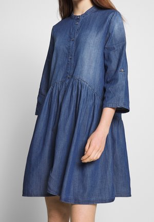 Žena v modrých džínových šatech s dlouhými rukávy, zapínáním na knoflíky vpředu, nařaseným pasem a rozšířenou sukní na jednoduchém světlém pozadí.