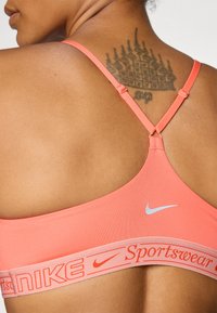 Nike sportski grudnjak u koraljnoj boji s tankim trakama, sadrži srebrni logo i elastičnu traku s natpisom "Nike Sportswear" u narančastoj boji.