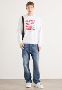 Felpa bianca con stampa grafica rossa, abbinata a jeans dritti azzurri e sneakers bianche. Design semplice e casual con una silhouette moderna.