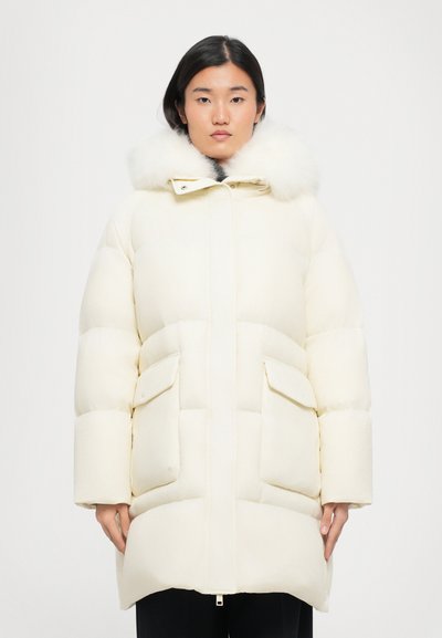 Puffjacke in Elfenbein mit gefütterter Kapuze aus Fell, horizontalem Steppmuster und zwei großen Fronttaschen. Reißverschluss vorne.