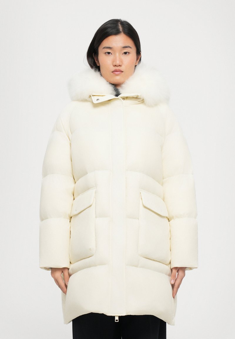 Puffjacke in Elfenbein mit gefütterter Kapuze aus Fell, horizontalem Steppmuster und zwei großen Fronttaschen. Reißverschluss vorne.