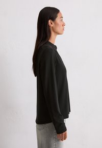 Chemise noire à manches longues avec col montant, texture lisse, coupe décontractée et fentes sur les côtes, portée avec un jean gris clair.
