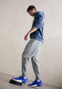 Sportsoutfit med en blå hættebluse og grå sweatpants. Sneakers er blå med hvide accenter og står på en sort vægtskive.
