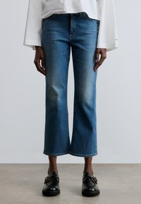 Kék farmer rövidített nadrág egyenes szárú, finom szegecselt részletekkel a szegély mentén. Laza fehér felsővel és fekete cipővel párosítva.