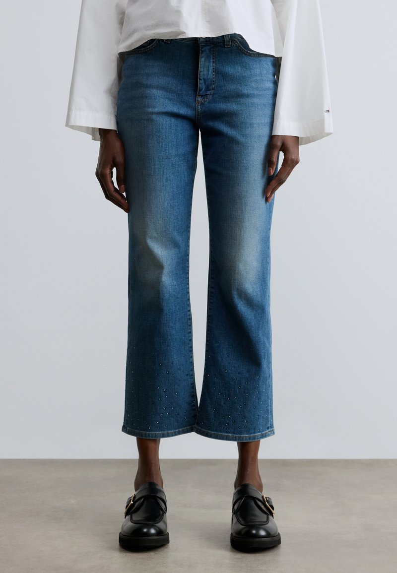 Kék farmer rövid�ített nadrág egyenes szárú, finom szegecselt részletekkel a szegély mentén. Laza fehér felsővel és fekete cipővel párosítva.