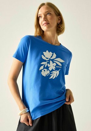 Kobieta w niebieskim t-shircie z białym kwiatowym wzorem i czarnej spódnicy, stoi z jedną ręką w kieszeni, a drugą trzyma brzeg koszulki.