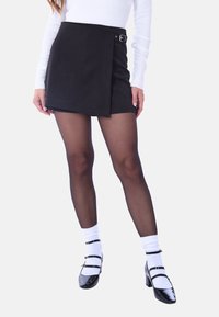 Gonna nera avvolgente con cintura, materiale texturizzato e orlo dritto. Abbinata a collant neri trasparenti e scarpe in pelle verniciata nera con cinturini.