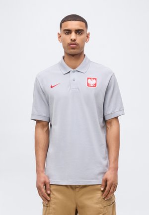 Jeune homme portant un polo gris clair avec un logo Nike rouge et un emblème d'aigle blanc, debout sur un fond clair uni.