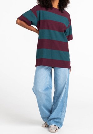 Persona che indossa una camicia a righe larga in rosso scuro e verde acqua con jeans azzurri chiari a gamba larga e sandali beige con punta chiusa, in piedi con una mano sui fianchi.