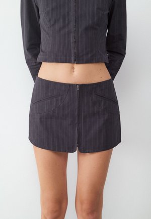 Mujer con mini falda oscura de rayas finas con cremallera frontal y chaqueta corta a juego, de pie frente a un fondo liso.