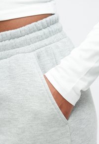 Mørkhudet person med hånd i lommen på lysegrå joggingbukser, iført en hvid langærmet top, der viser maven og detaljer med elastisk taljebånd.