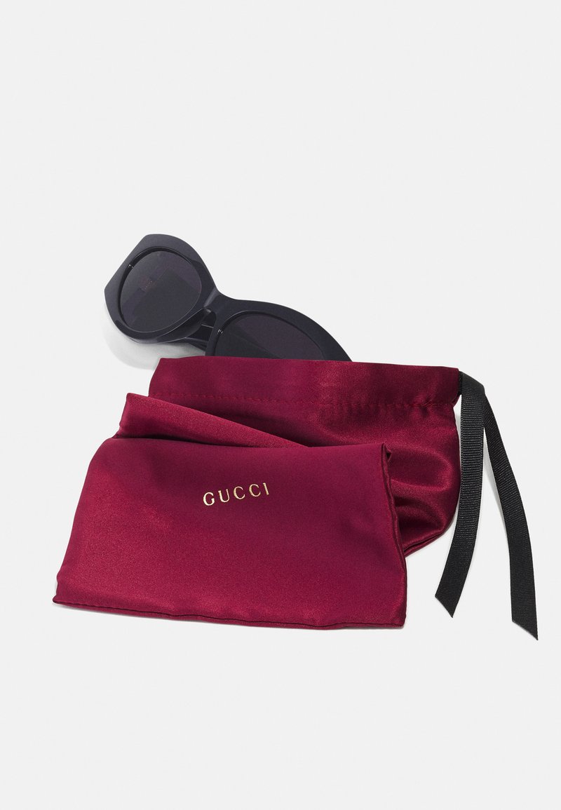 Gucci schwarze Sonnenbrille mit rechteckigem Rahmen, liegend auf einem bordeauxroten Satinbeutel mit goldgeprägtem Logo und schwarzen Bandbindungen.