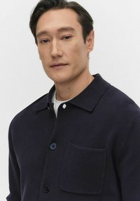 Mörkblå stickad skjortjacka med krage, knäppning med knappar och en bröstficka. Slät textur med en strukturerad design.