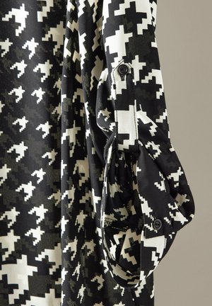 Mustavalkoinen kuviollinen kangas, jossa on sahalaitakuvio. Pitkähiha, jossa napitusyksityiskohta, sileä pinta. Läheltä otettu kuva vaatteesta.