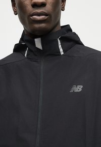 Κοντινό πλάνο ενός ατόμου που φοράει μαύρο μπουφάν με φερμουάρ και κουκούλα, με αντανακλαστικές λωρίδες και λογότυπο New Balance στο στήθος.