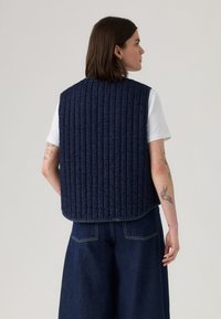 Gestepptes navyfarbiges Weste aus strapazierfähigem Stoff, mit vertikalen Streifen, ärmellosem Design und abgerundetem Saum, über einem weißen T-Shirt getragen.