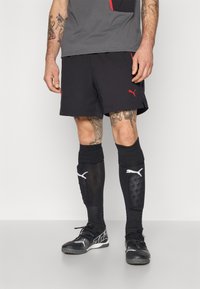 Sorte atletiske shorts med røde accenter, parret med sorte skinnebeskyttere med hvidt logo. Sorte sportssko med struktureret design.