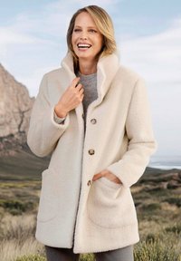 Fleece jas in crème met een hoge kraag, sluiting met knopen aan de voorkant en zijzakken; heeft een zachte textuur en afgeronde zomen.