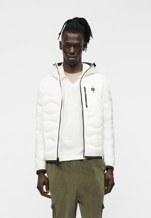 Tânăr purtând o geacă albă cu fermoar peste un pulover deschis la culoare și pantaloni din catifea cu nervuri verde măsliniu, stând pe un fundal simplu.