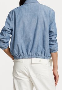 Jasnoniebieska kurtka dżinsowa z marszczonym dołem i podwiniętymi rękawami. Tył ma klasyczny kołnierz i minimalistyczny design. Noszona na białych spodniach.