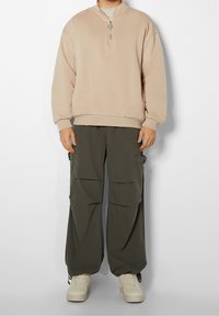Sudadera beige con cremallera y cuello alto, combinada con pantalones cargo holgados en color oliva que presentan varios bolsillos y un acabado texturizado.