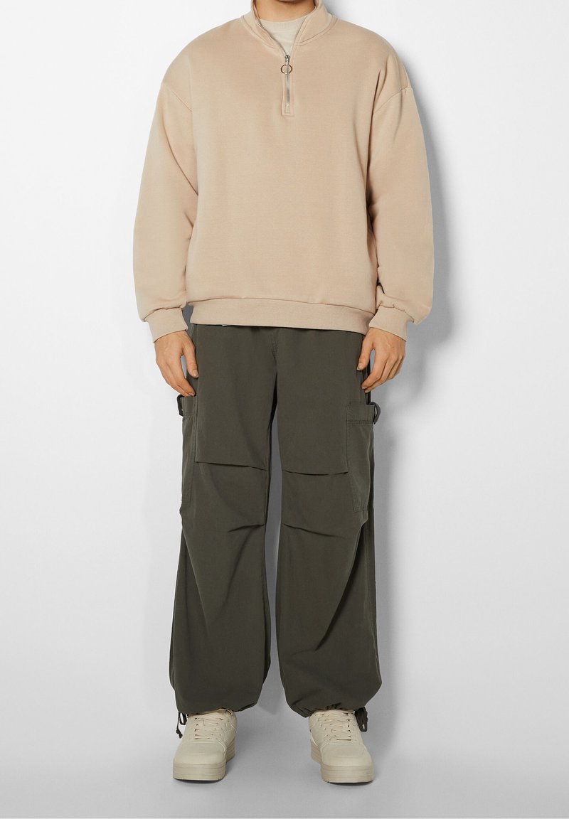 Sudadera beige con cremallera y cuello alto, combinada con pantalones cargo holgados en color oliva que presentan varios bolsillos y un acabado texturizado.