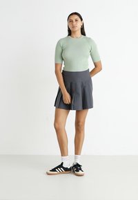 Haut vert clair à rayures, à manches courtes, jupe plissée noire, baskets noires avec des rayures blanches, portées avec des chaussettes blanches, debout sur un fond uni.