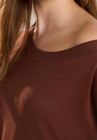 Brun off-shoulder striktrøje med en tekstureret overflade og ribbet halsudskæring. Det bløde stof falder let og fremhæver en strømlinet silhuet.
