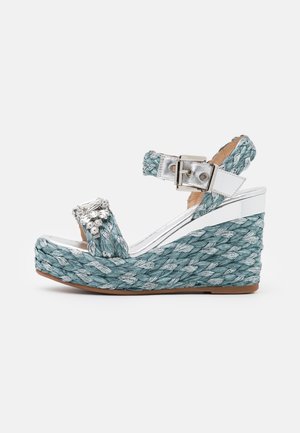 Wedge sandal med en vævet teal overdel, sølv accenter og en rem med en spænde. Platformshøjden forbedrer holdbarheden og stabiliteten.