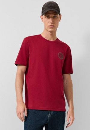 Jonge man die een rood T-shirt met korte mouwen en klein borstlogo draagt, donkere spijkerbroek, zwarte pet en kleine oorbellen, staand tegen een effen achtergrond.