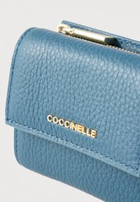 Portafoglio in pelle blu con texture, dotato di zip dorata e nome del marchio "COCCINELLE" in oro sul risvolto anteriore, su uno sfondo bianco.