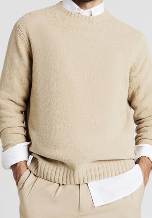 Mand iført en beige strikket sweater over en hvid skjorte med beige bukser, hænderne i lommerne, mod en ensfarvet baggrund.