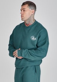 Sudadera de color verde azulado con cuello redondo, mangas largas y puños acanalados. Presenta un pequeño logotipo blanco en el pecho y un corte relajado.
