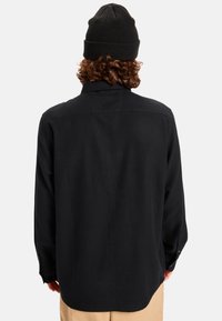Zwarte langslagen shirt met een ontspannen pasvorm en knoopmanchetten, met een gladde textuur en ronde zoom. Draagt met een zwarte beanie.