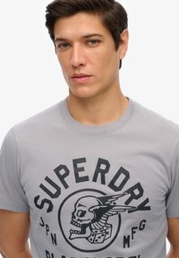 Grijze katoenen t-shirt met een grafisch ontwerp van een schedel en dobbelstenen, met de tekst "Superdry" in zwart. Ronde hals en korte mouwen. Gladde textuur.
